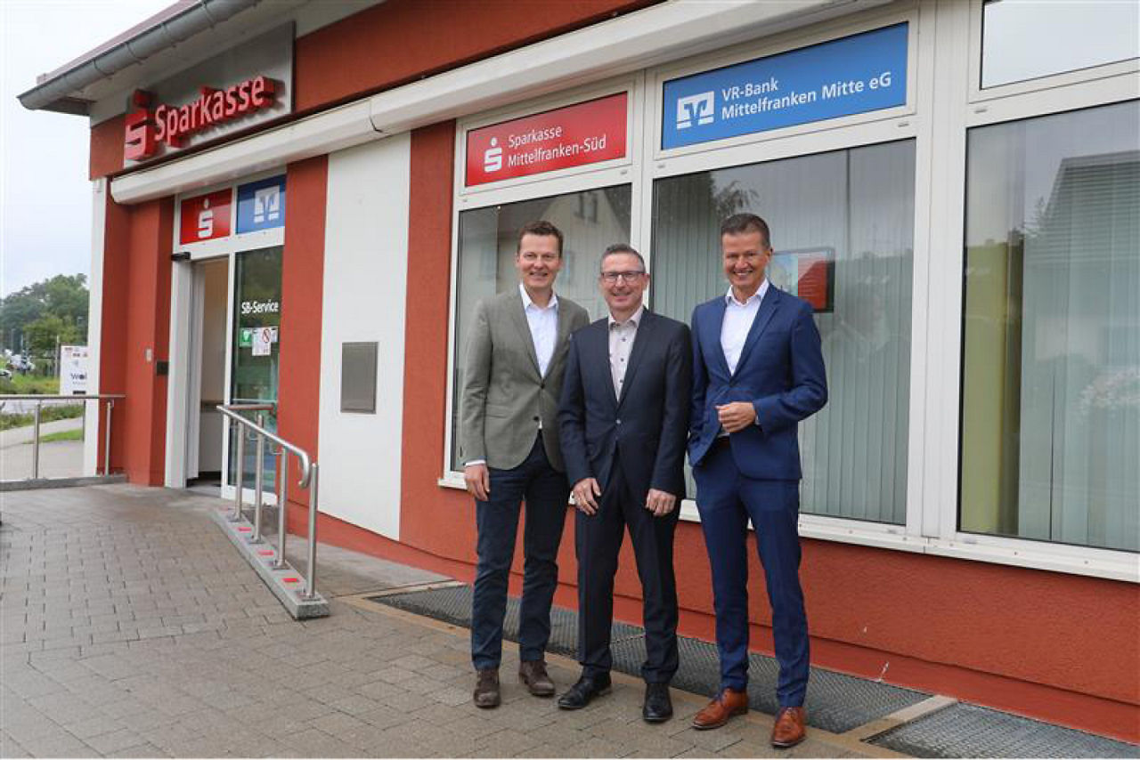 Drei Personen stehen nebeneinander und lächeln, sie stehen vor der gemeinsamen Filiale mit der Sparkasse