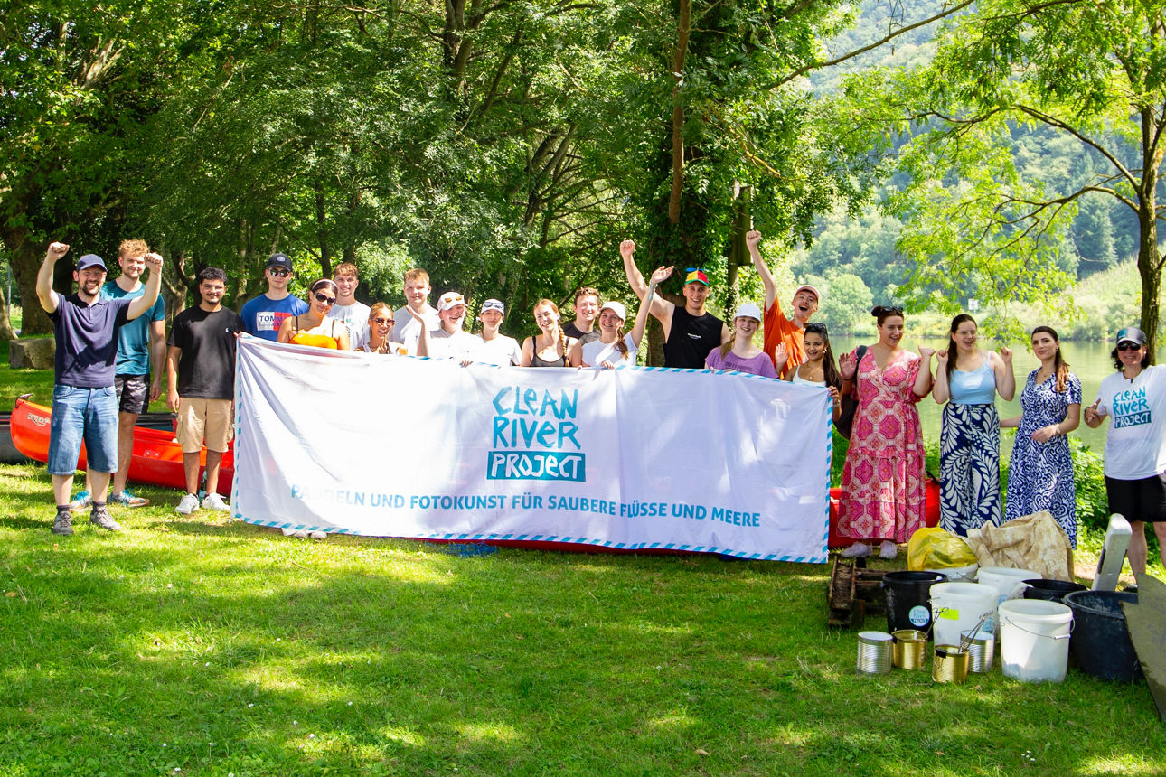 Gruppe mit Banner „Clean River Project – Paddeln und Fotokunst für saubere Flüsse und Meere“, Kanus und gesammeltem Müll am F