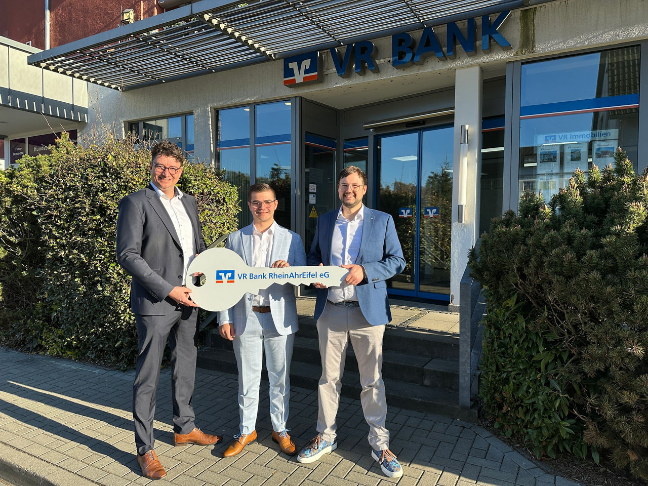 Drei Personen stehen vor einer Bankfiliale und halten gemeinsam einen großen Schlüssel in den Händen.