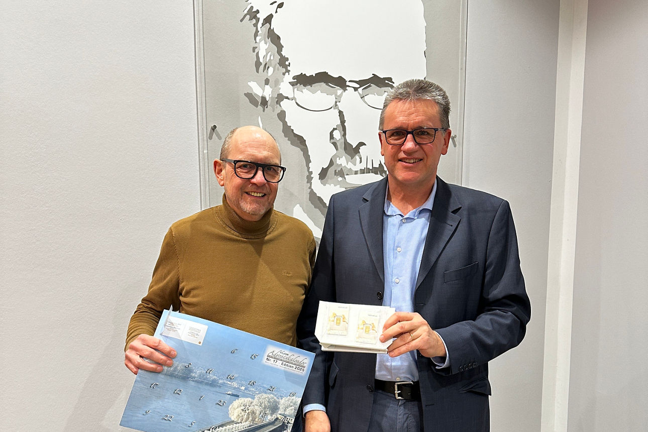 Zwei Personen präsentieren Briefmarken und ein Poster vor Wandkunst bei einer offiziellen Veranstaltung.