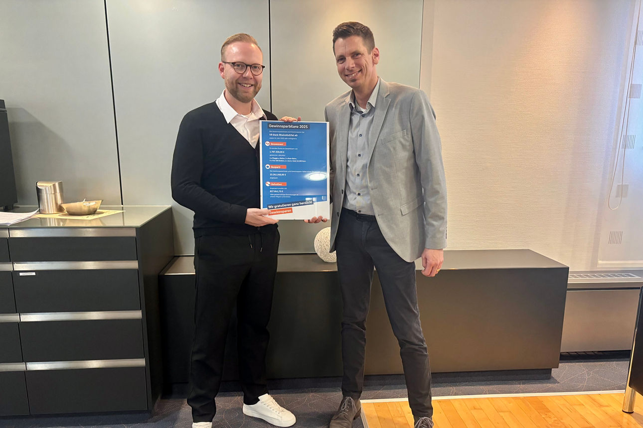 Zwei Personen halten im Büro ein Poster „Gewinnspielkarte 2025“ mit fünf Aktionen und „Wir gratulieren ganz herzlich!“