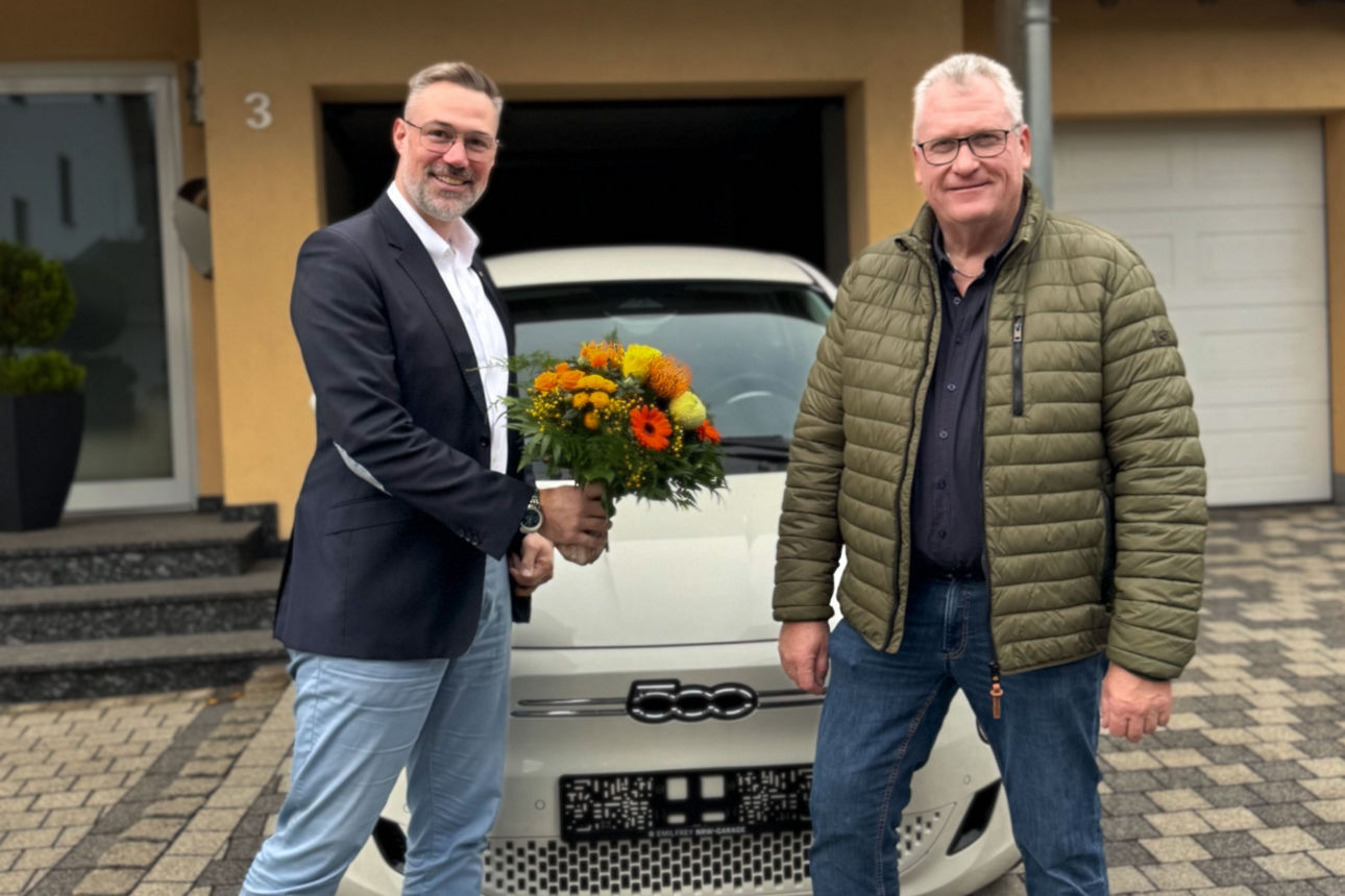 Zwei Männer bei der Gewinnübergabe eines Fiat 500 vor Haus Nr. 3, einer hält einen Blumenstrauß.