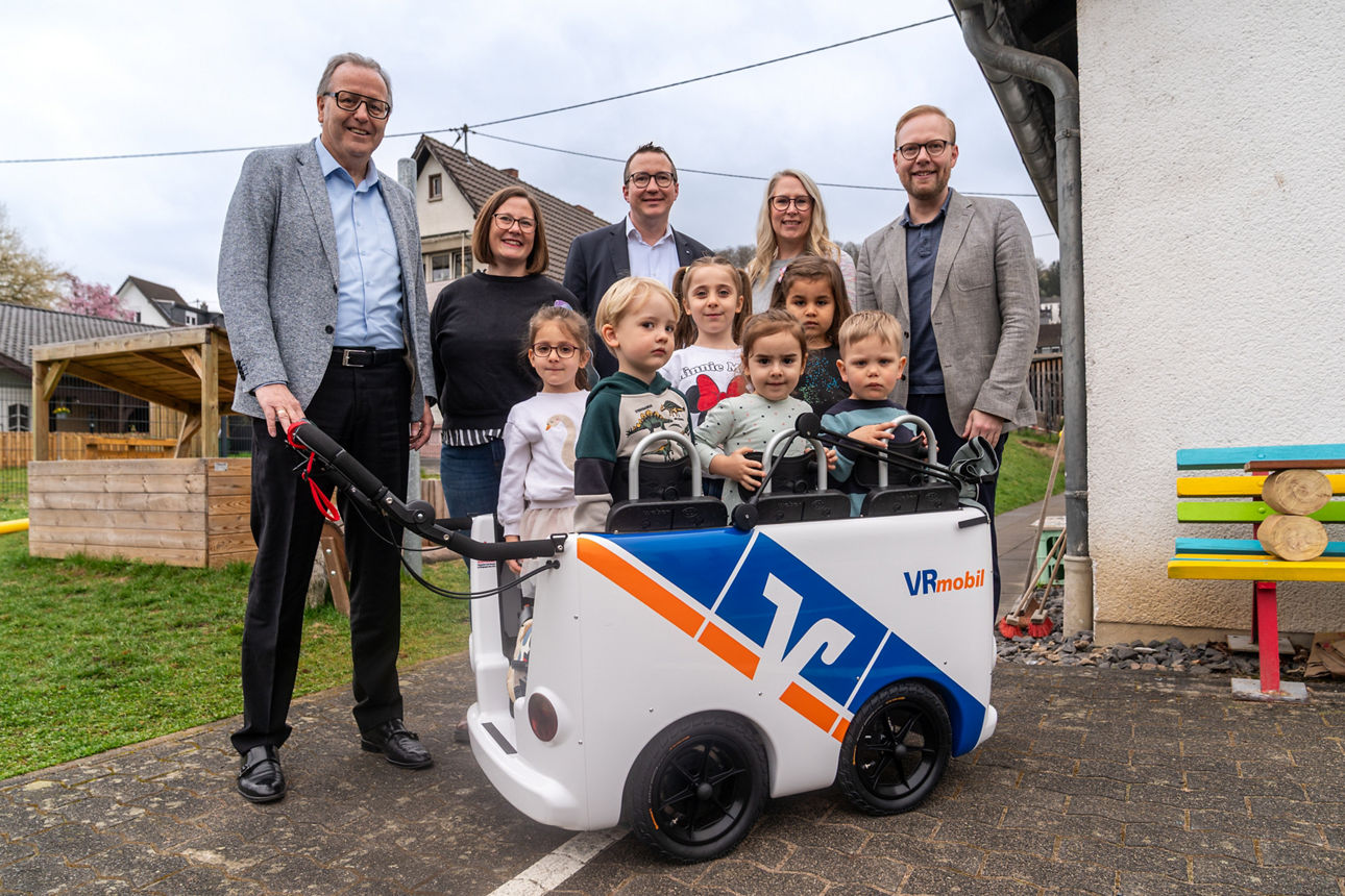 Mehrere Erwachsene stehen auf einem Spielplatz hinter Kindern, die in einem Kinderbus sitzen.