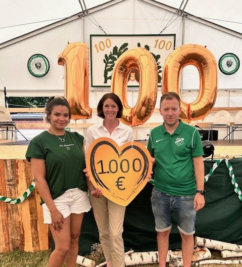 Zwei Frauen und ein Mann halten einen Spendenscheck in Höhe von 1000 Euro in Herzform. Im Hintergrund ist ein Festzelt und die Zahl 100 aus Luftballons zu sehen.
