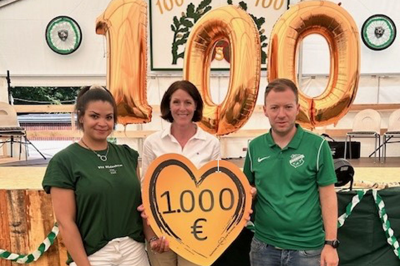 Zwei Frauen und ein Mann halten einen Spendenscheck in Höhe von 1000 Euro in Herzform. Im Hintergrund ist ein Festzelt und die Zahl 100 aus Luftballons zu sehen.