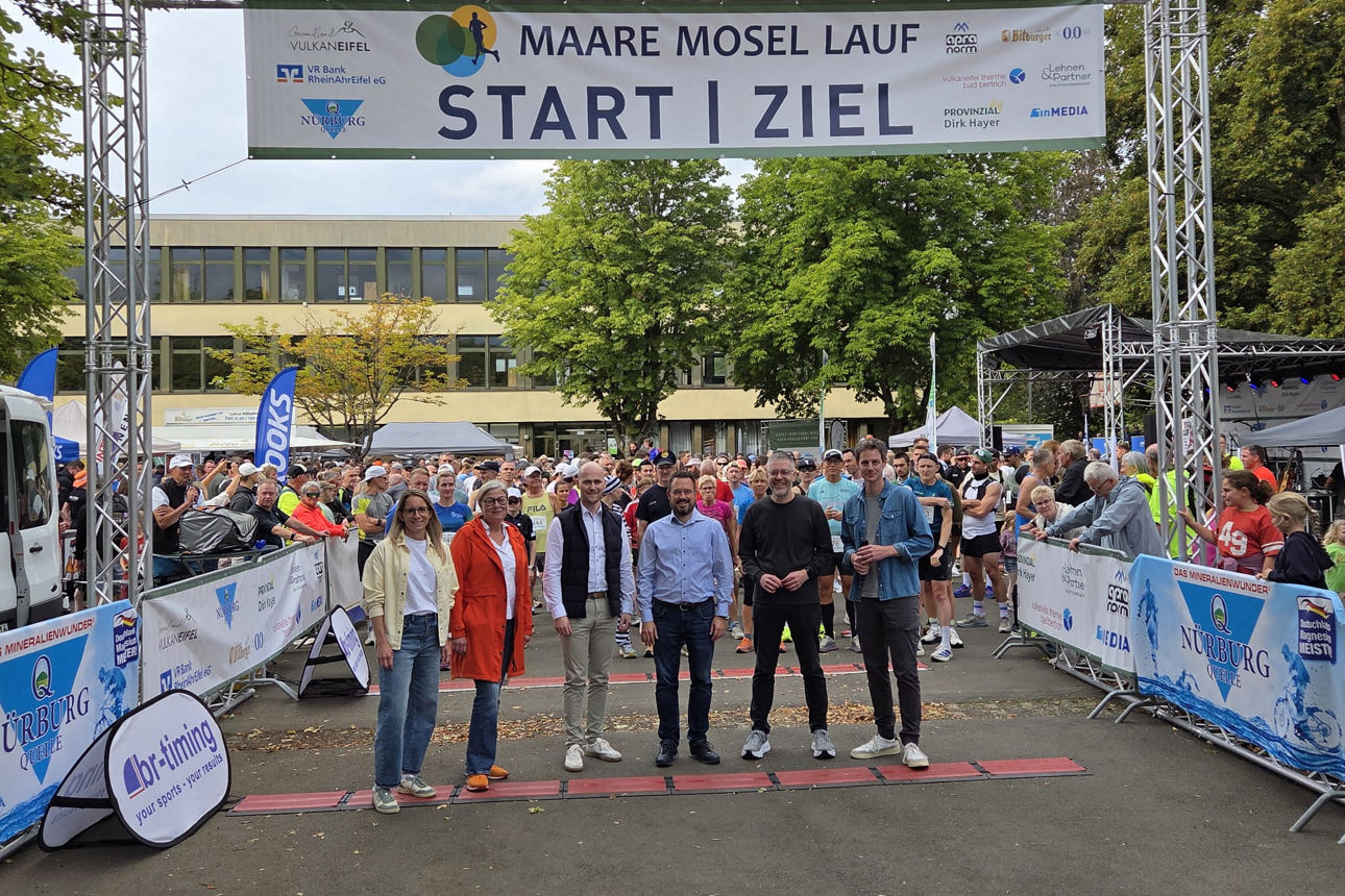 Gruppe von Läuferinnen und Läufern am Startbereich des Maare Mosel Laufs vor einem Banner mit Sponsorenlogos, darunter die VR