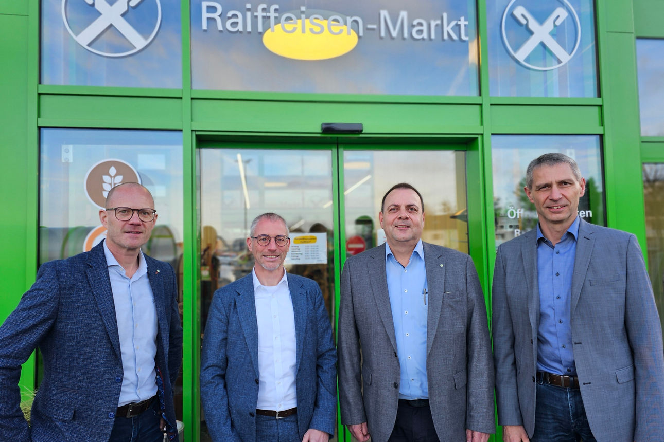 Vier Männer im Business-Outfit vor dem Eingang eines Raiffeisen-Markts mit grünem Rahmen und Schriftzug.