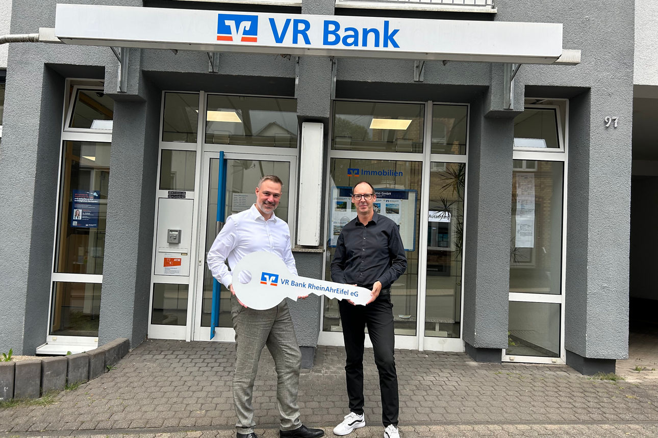 Zwei Männer halten einen großen Schlüssel vor dem Eingang einer VR Bank-Filiale.