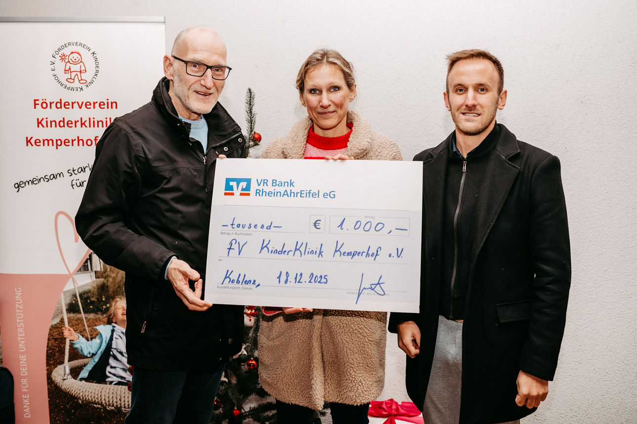 Drei Personen übergeben symbolischen Scheck der VR Bank RheinAhrEifel eG über 1.000 € an den Förderverein Kinderklinik Kemper