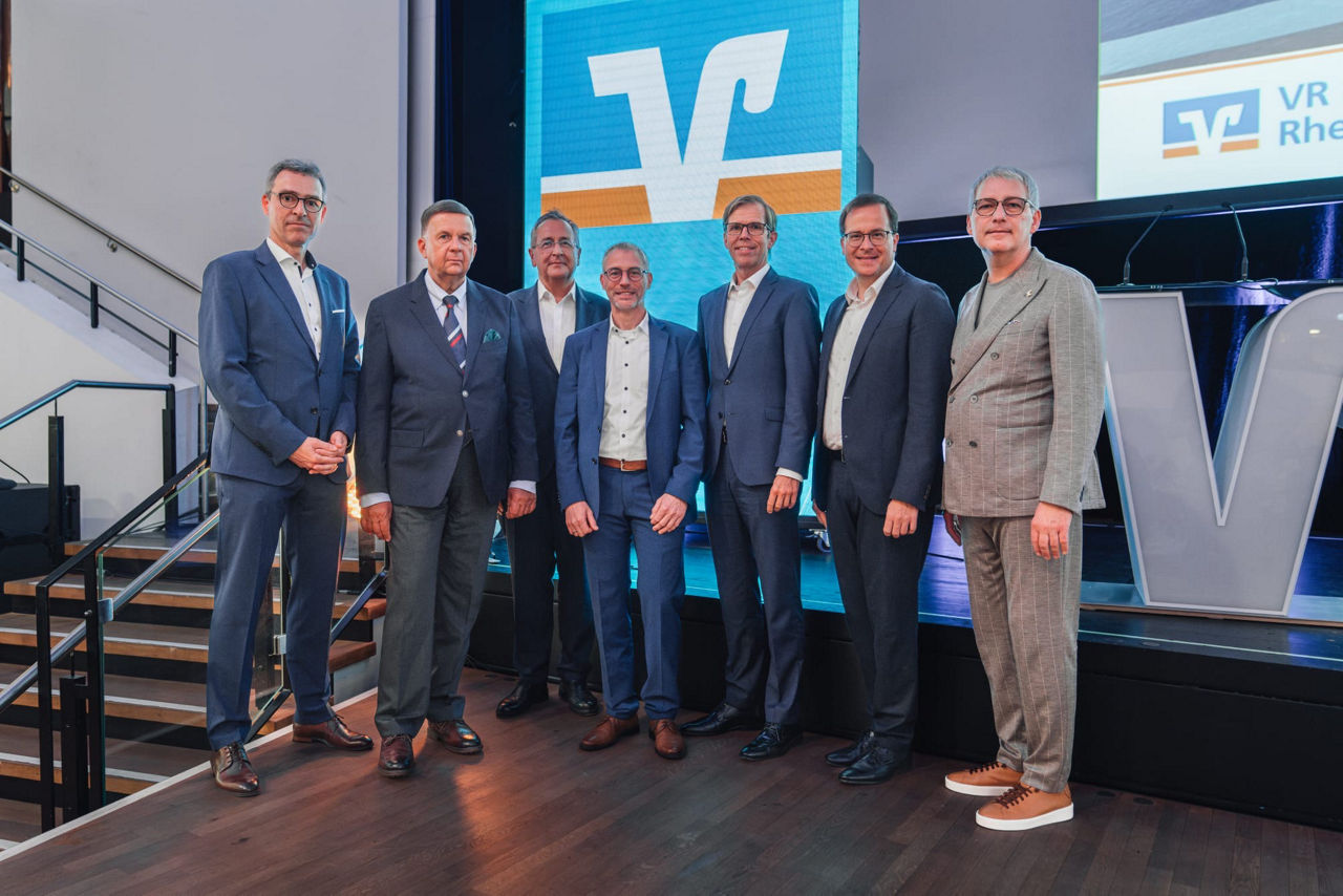 Mehrere Personen stehen vor einer Videowand, die das Markenzeichen der VR Bank zeigt, zu einem Gruppenbild zusammen.