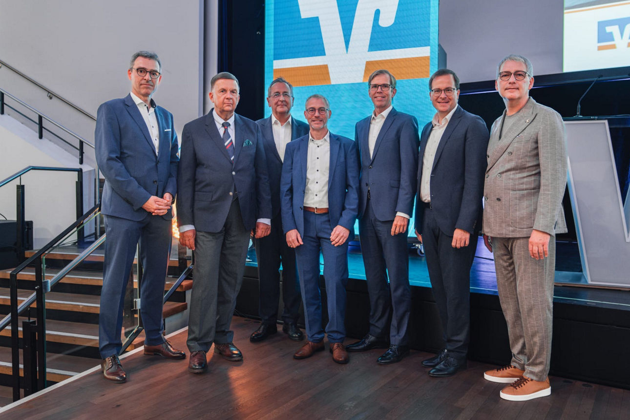 Mehrere Personen stehen vor einer Videowand, die das Markenzeichen der VR Bank zeigt, zu einem Gruppenbild zusammen.