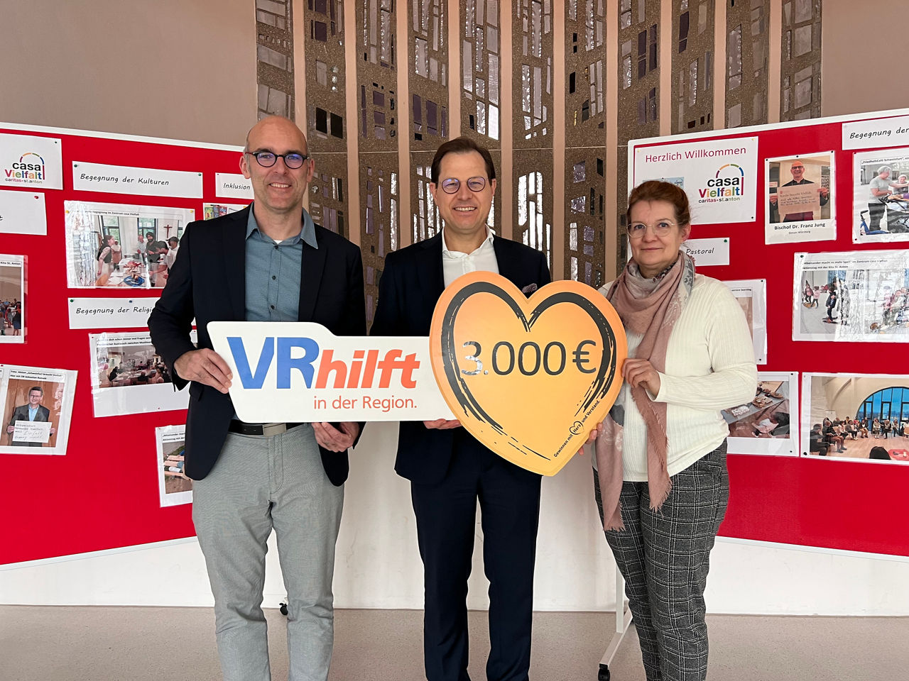 Drei Personen stehen gemeinsam vor einer Infowand und halten ein Schild mit dem Spendenbeitrag von 3.000 €.
