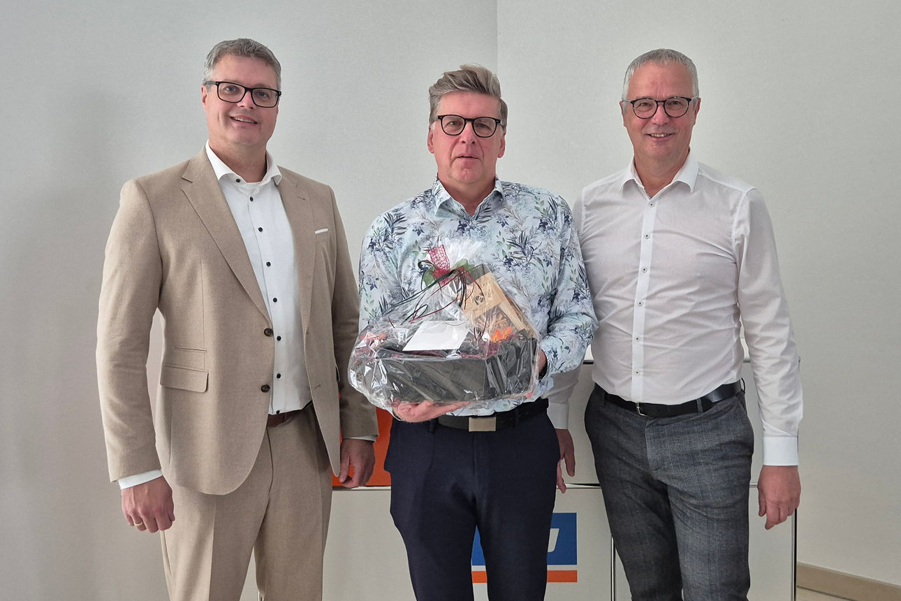 Drei Männer stehen nebeneinander in einem hellen Raum. Der Mann in der Mitte hält einen Geschenkkorb in den Händen. 