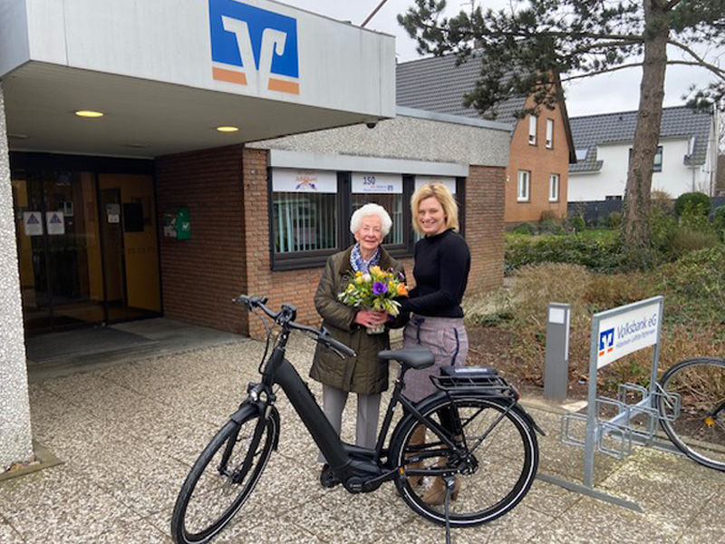 Eine ältere Dame mit Blumenstrauß in der Hand und eine Volksbank-Mitarbeiterin aus Arpke stehen lächelnd hinter einem E-Bike.
