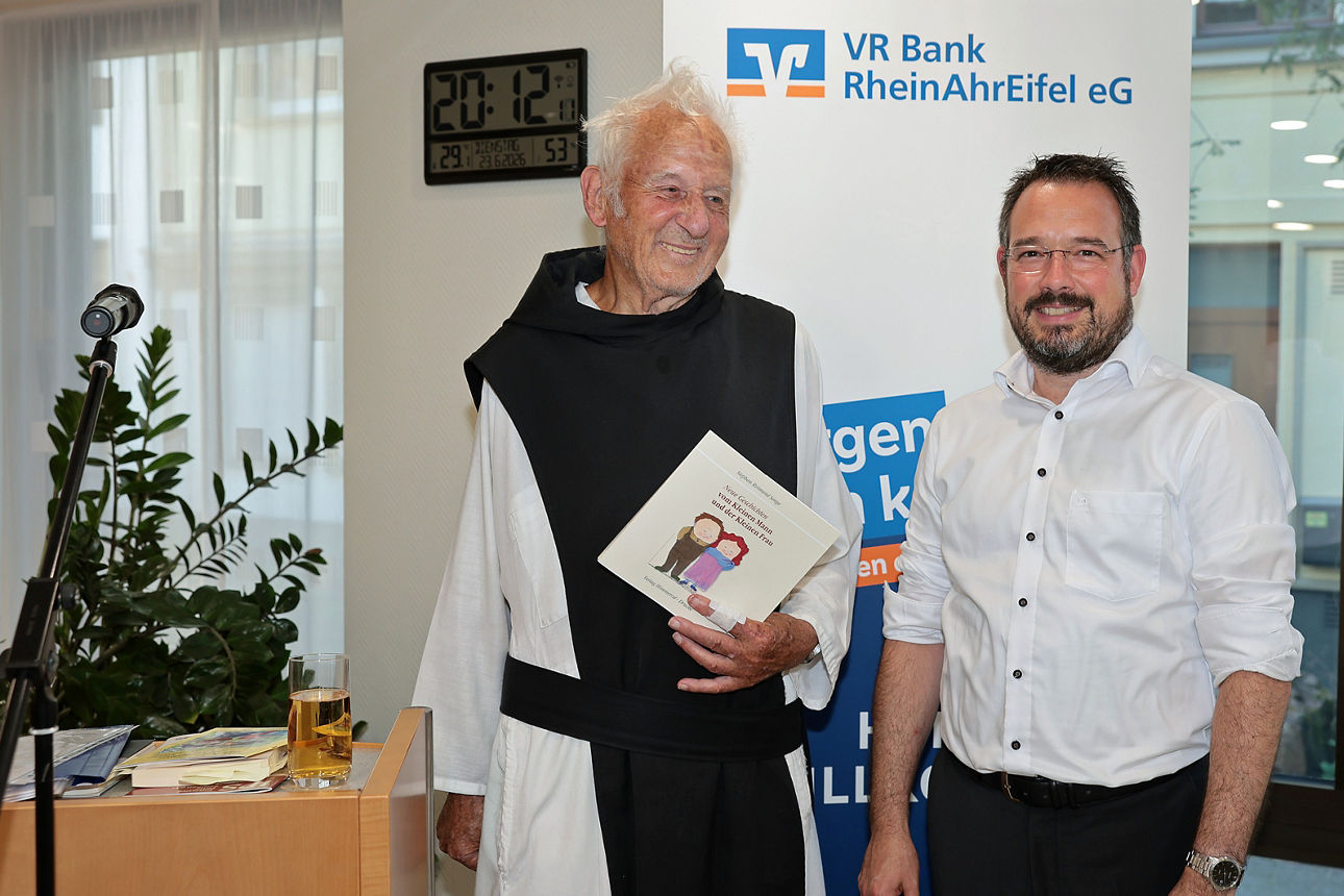 Zwei Männer bei der VR Bank RheinAhrEifel eG in Adenau; der ältere hält ein illustriertes Buch, Mikrofon am Tisch.