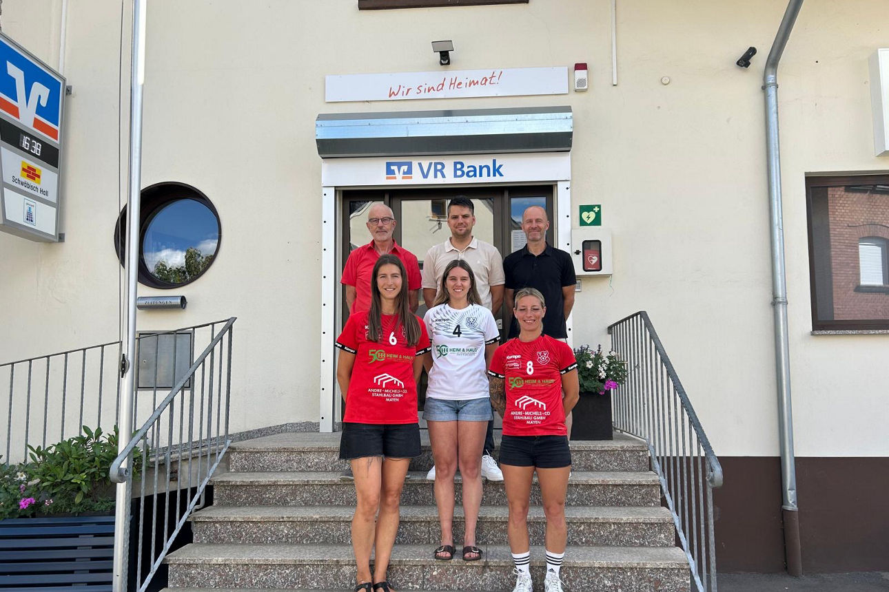 Drei Handballerinnen in roten und weißen Sporttrikots posieren gemeinsam mit Vereins- und Bankvertretern für ein Gruppenfoto