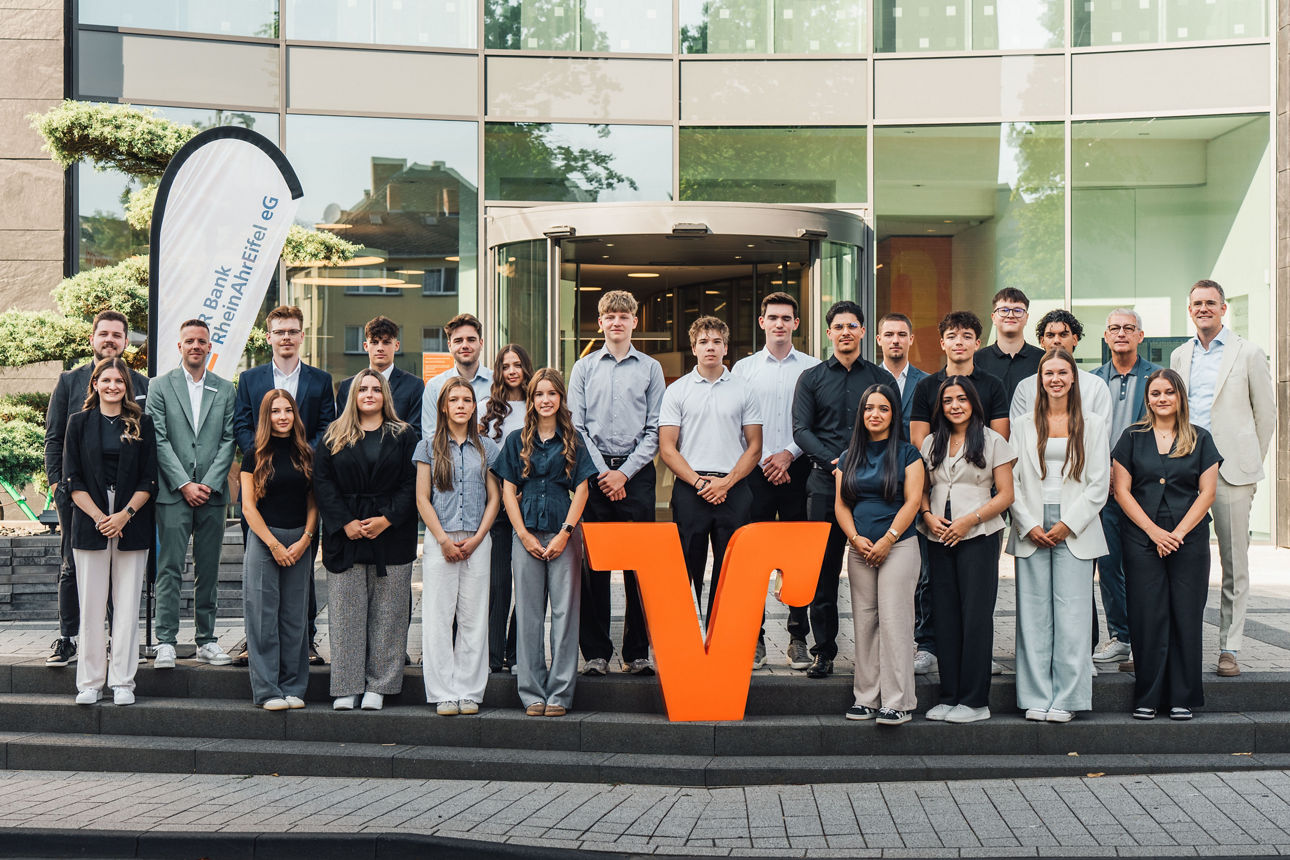Mitarbeitende der VR Bank RheinAhrEifel eG vor modernem Bankgebäude, gruppiert um großes oranges V-Logo