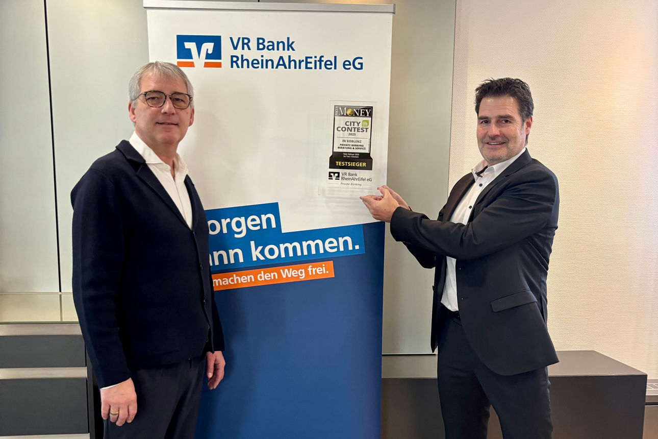 Zwei Personen stehen in einem Raum vor einem Roll-Up und präsentieren eine Trophäe.