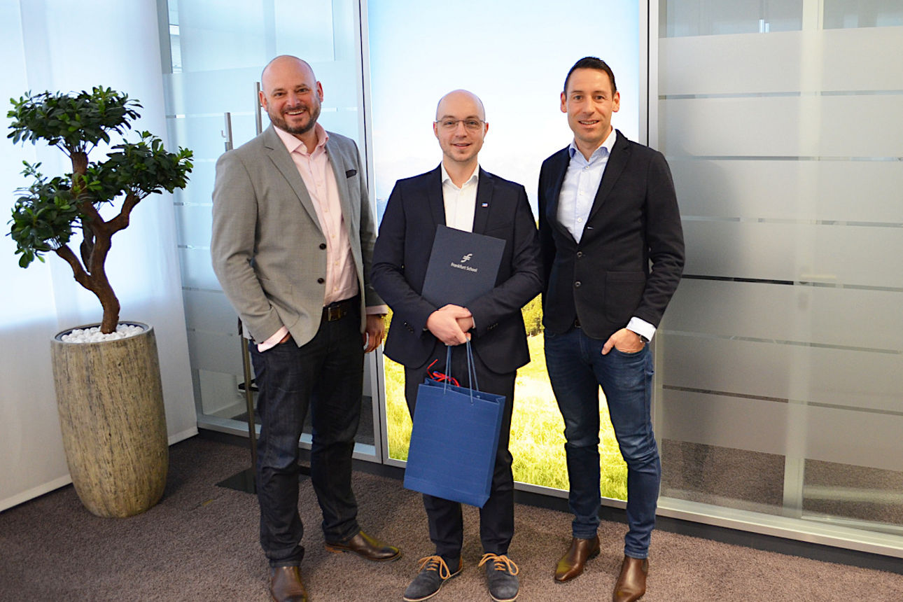 Mitarbeiter steht mit Urkunde und Geschenkkorb neben beiden Vorständen nebeneinander in der Filiale vor einer Glastrennwand