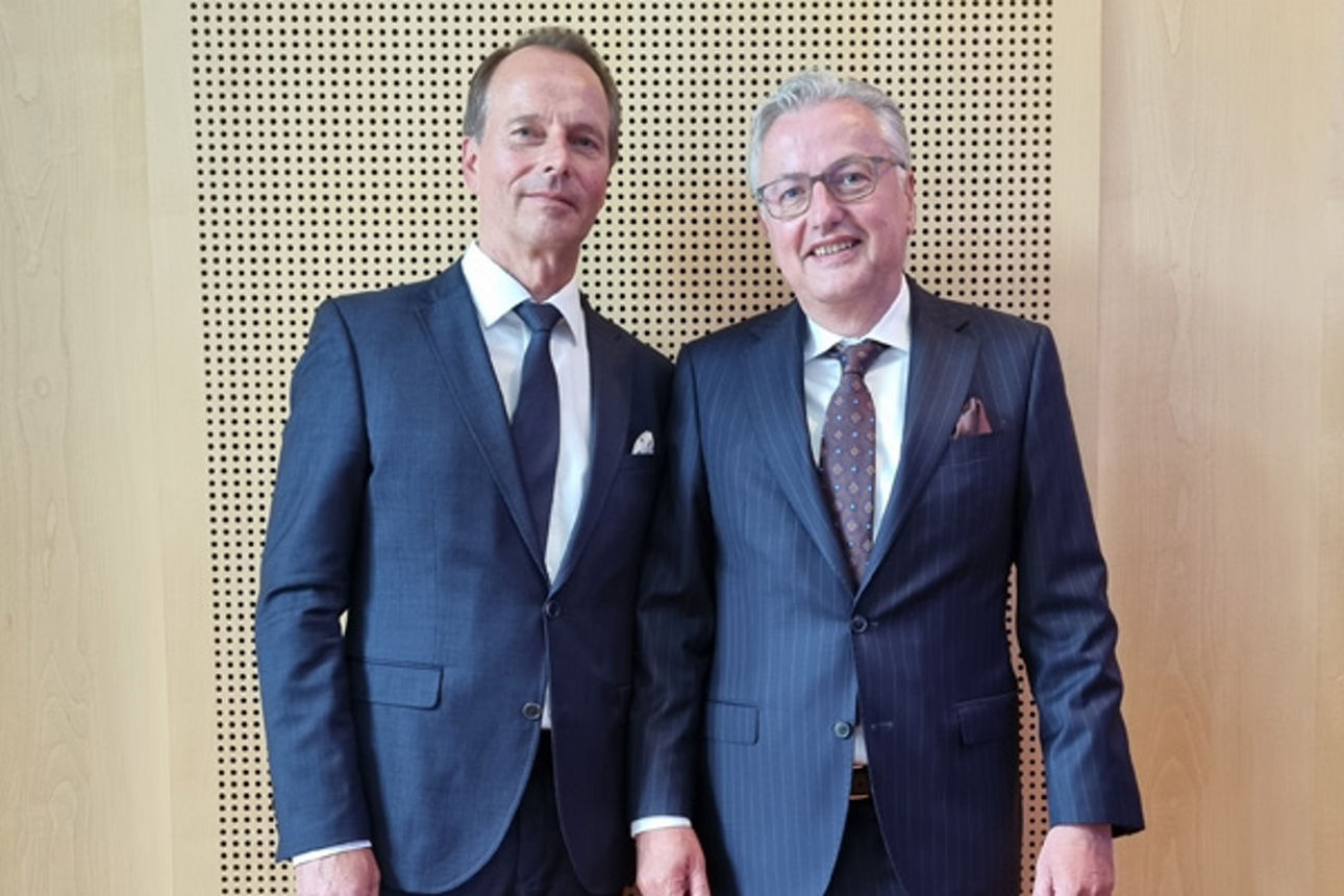 Zwei Männer im Anzug stehen vor einer beige-braunen Wand.