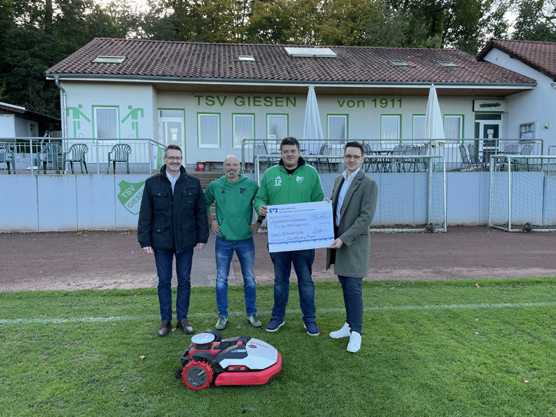Vier Männer stehen hinter einem Mähroboter auf dem Fußballplatz und halten einen Überreichungsscheck in der Hand.