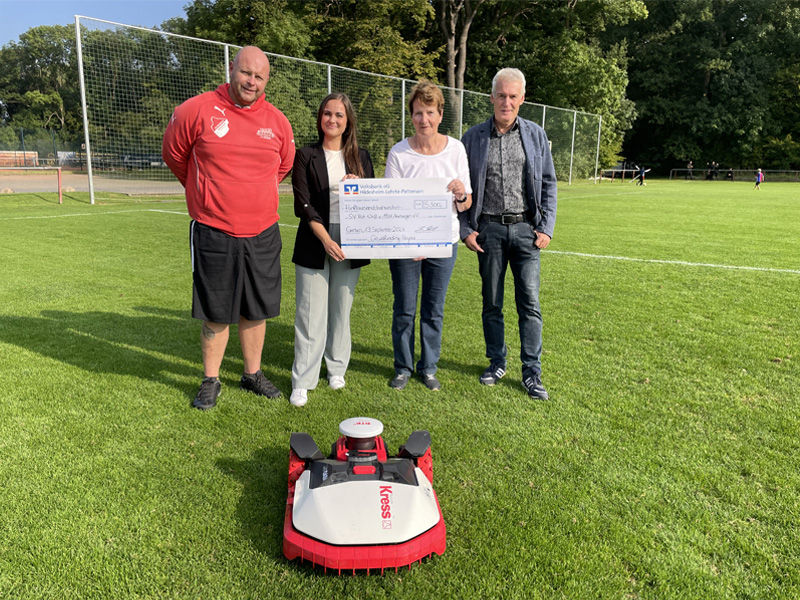 Zwei Männer und zwei Frauen stehen hinter einem Mähroboter und halten einen Überreichungsscheck in der Hand.