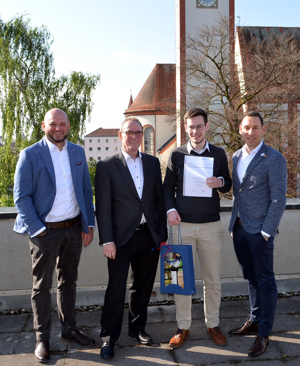 Mitarbeiter steht mit Urkunde und Geschenkkorb mit drei Führungskräften auf der Dachterrasse mit Kirche im Hintergrund