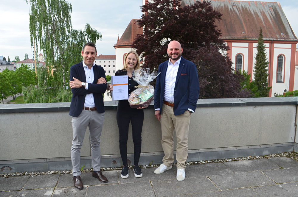 Mitarbeiterin steht mit Urkunde und Geschenkkorb neben den beiden Vorständen auf einer Dachterrasse
