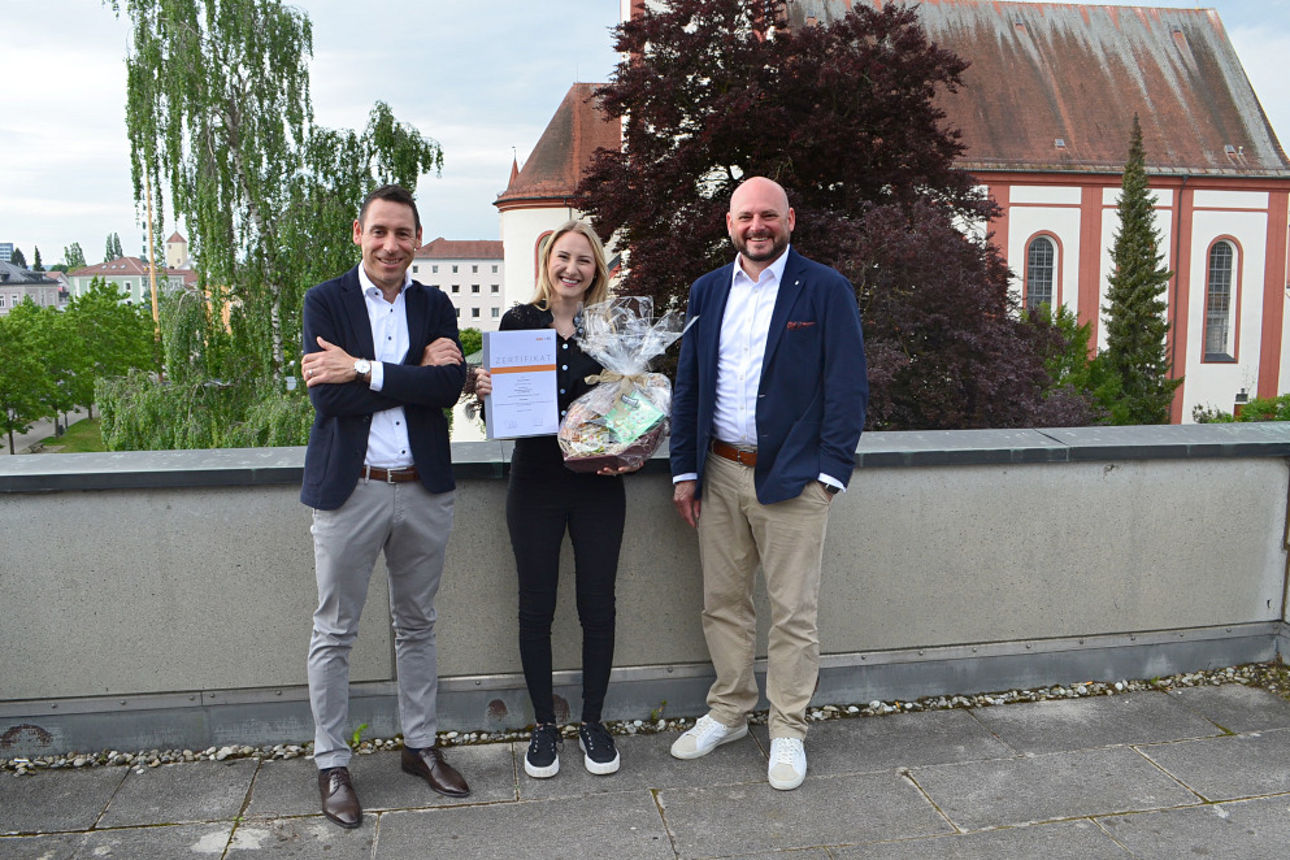 Mitarbeiterin der Bank steht mit Urkunde neben zwei Vorständen auf einer Dachterasse