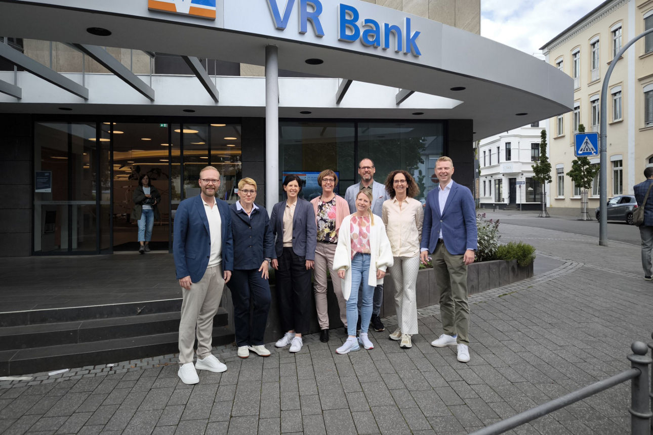 Acht Mitarbeitende der VR Bank RheinAhrEifel eG in Business-Kleidung vor dem Eingang einer Filiale mit VR-Logo