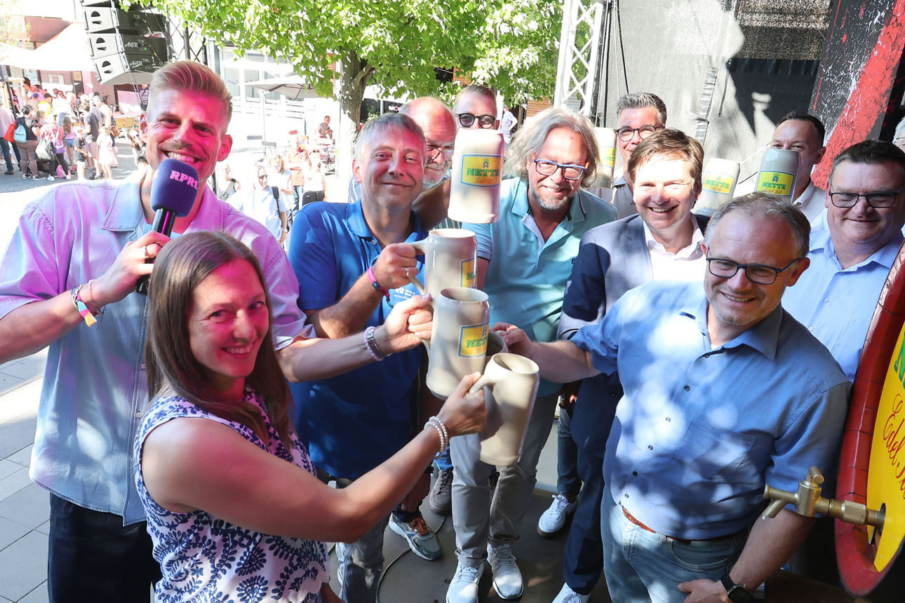 Gruppe stößt beim Deichstadtfest mit Keramikbierkrügen an; Moderator hält RPR1-Mikrofon.