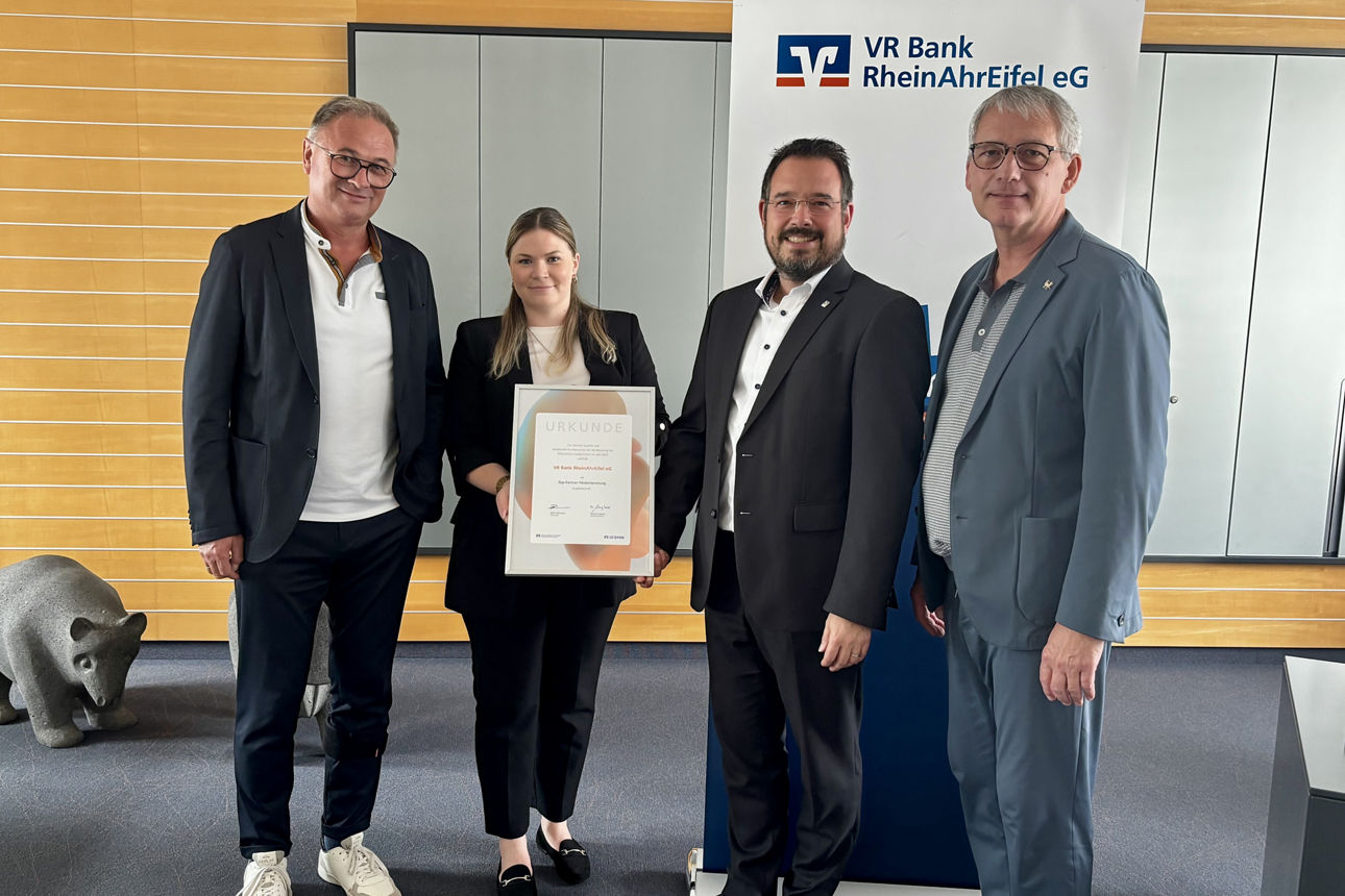 Vier Personen bei VR Bank RheinAhrEifel eG; Frau hält gerahmte Urkunde, Banner mit Banklogo im Hintergrund.