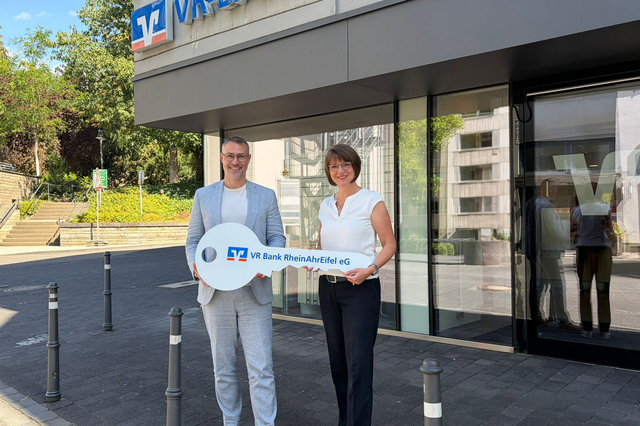 Zwei Filialleitende halten vor der VR Bank-Filiale ein rundes Schild „VR Bank RheinAhrEifel eG