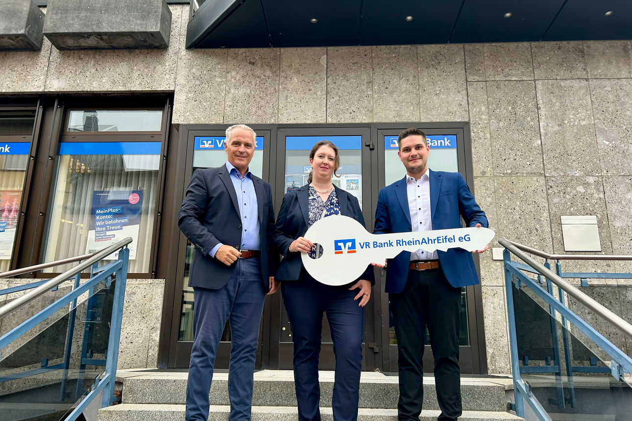 Drei Personen stehen vor einer Bankfiliale und halten gemeinsam einen großen symbolischen Schlüssel in der Hand.