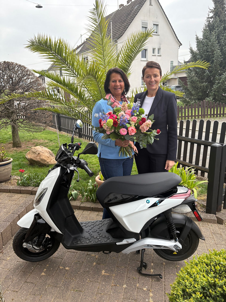 Zwei Personen stehen hinter einem E-Roller. Eine Frau hat einen Blumenstrauß in der Hand.