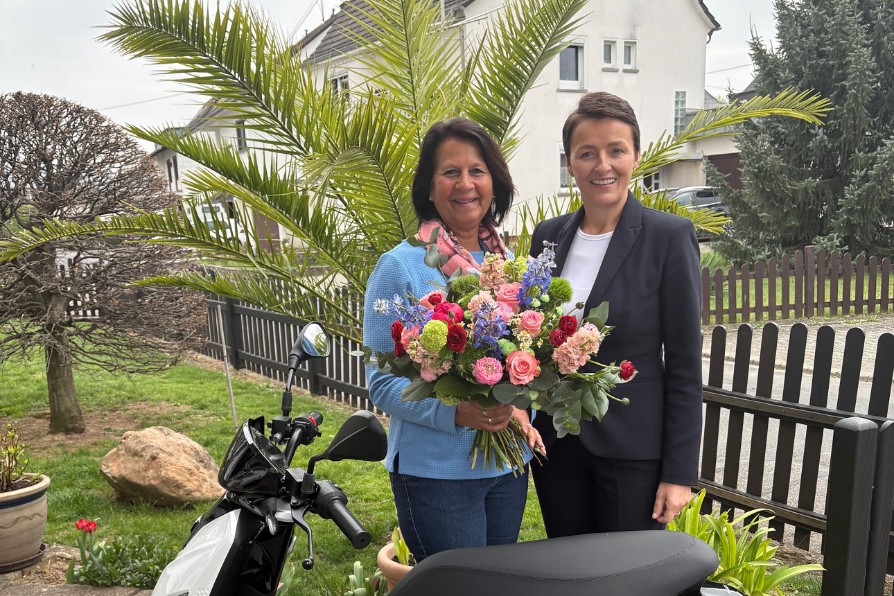Zwei Personen stehen hinter einem E-Roller. Eine Frau hat einen Blumenstrauß in der Hand.