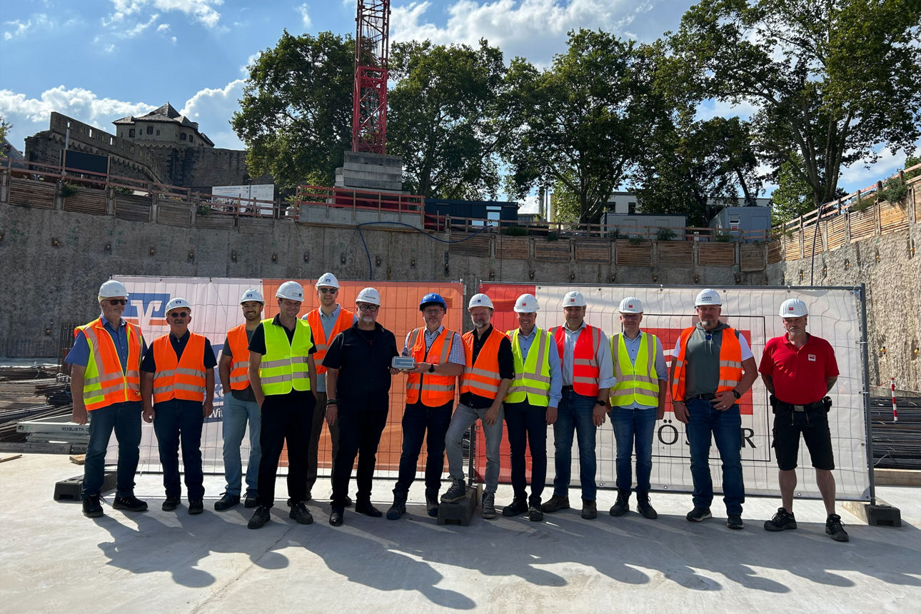 13 Personen mit Helmen und Warnwesten bei Grundsteinlegung auf Baustelle Andernach, Kran und Burg im Hintergrund.