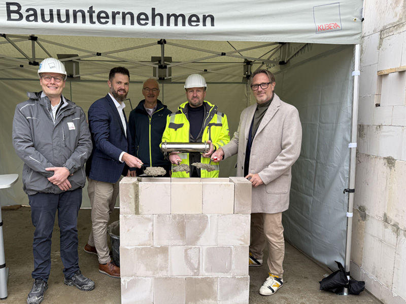 Fünf Männer stehen hinter einem Stück Mauer, ein Mann mit Bauhelm hält eine Zeitkapsel in der Hand