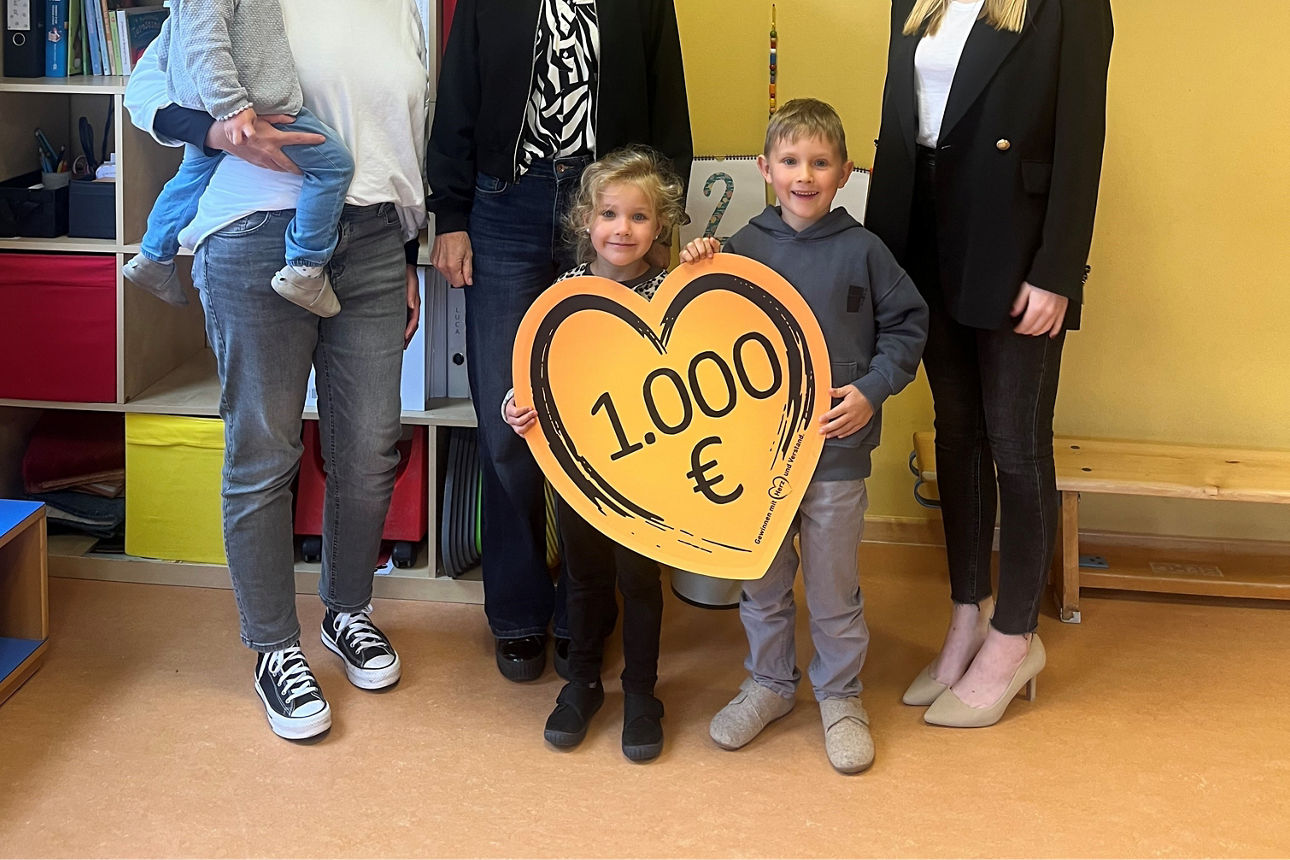 Drei Frauen und drei Kinder stehen vor einer gelben Wand. Die beiden Kinder in der Mitte halten ein Herz mit der Spendensumme von 1.000 Euro nach oben.