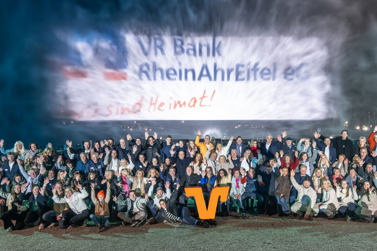 Gruppe Menschen posiert abends vor beleuchtetem „VR Bank RheinAhrEifel eG. Es sind Heimat!“-Schriftzug bei Klangwelle.