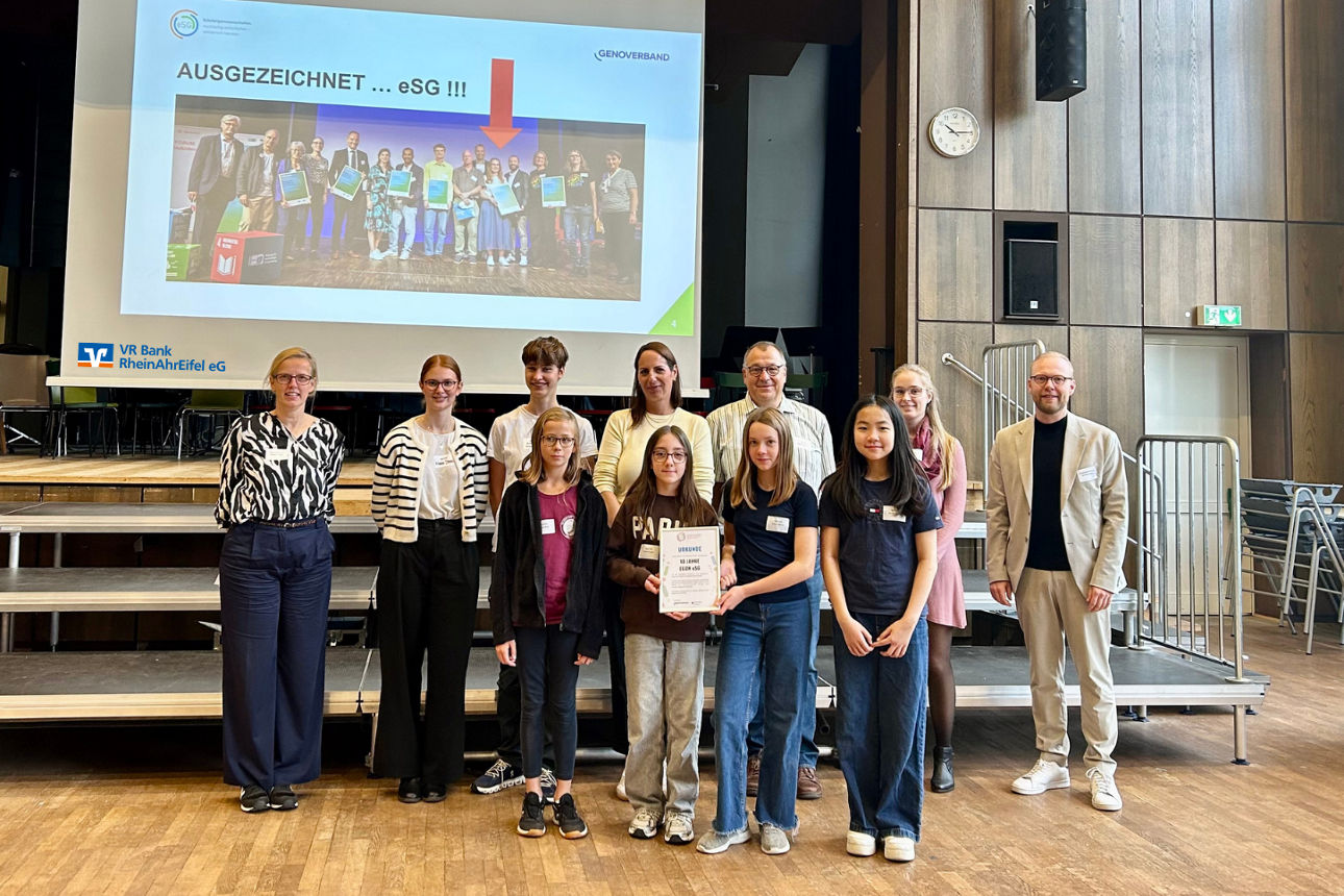 Gruppe von Schüler:innen und Erwachsenen auf Bühne, Schülerin hält Urkunde, Projektion: „AUSGEZEICHNET ... eSG !!!“
