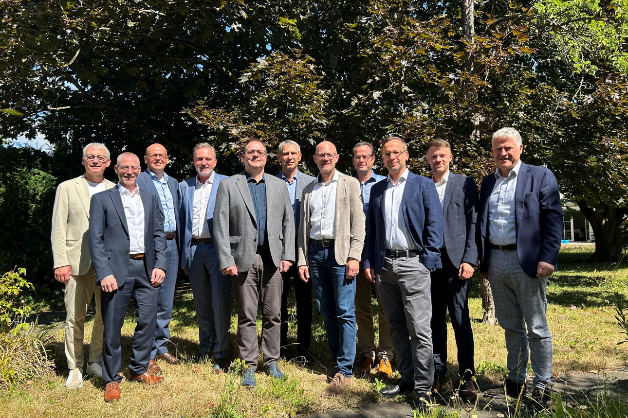 Elf Männer in Businesskleidung stehen für ein Pressefoto auf einer Wiese vor dichtem grünen Baumbestand