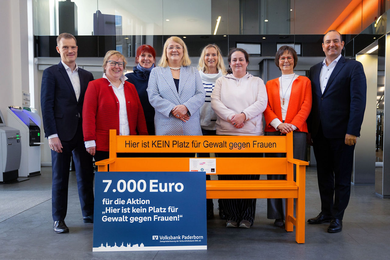 Eine kleine Gruppe steht hinter einer orangen Bank und einem Spendenscheck über 7.000 Euro