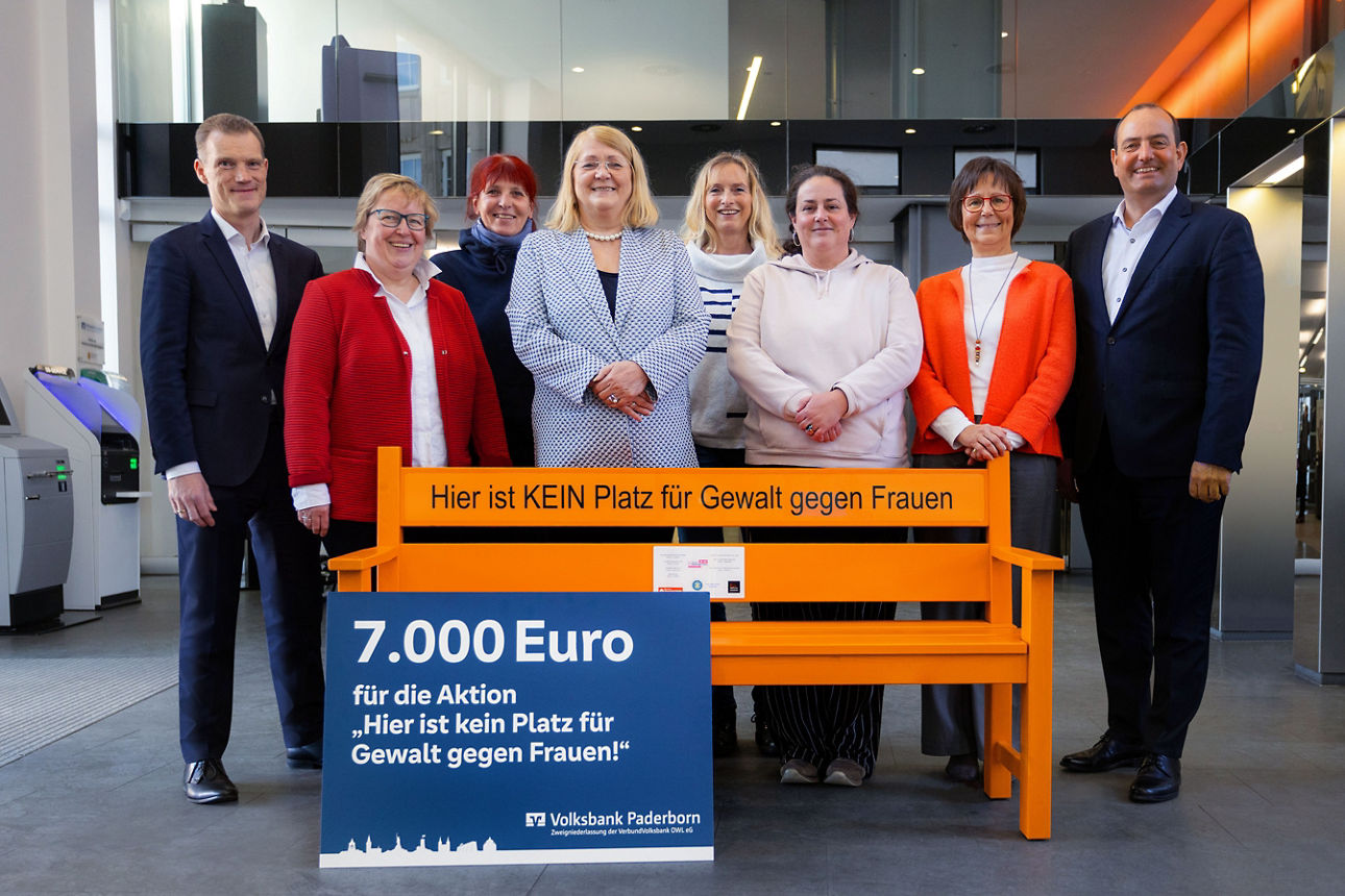 Eine kleine Gruppe steht hinter einer orangen Bank und einem Spendenscheck über 7.000 Euro