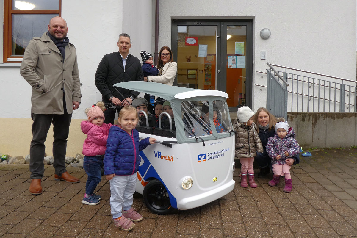 Kinder stehen mit Kinderbus und Mitarbeitern der Bank mit der Kindergartenleitung vor dem Kindergarten