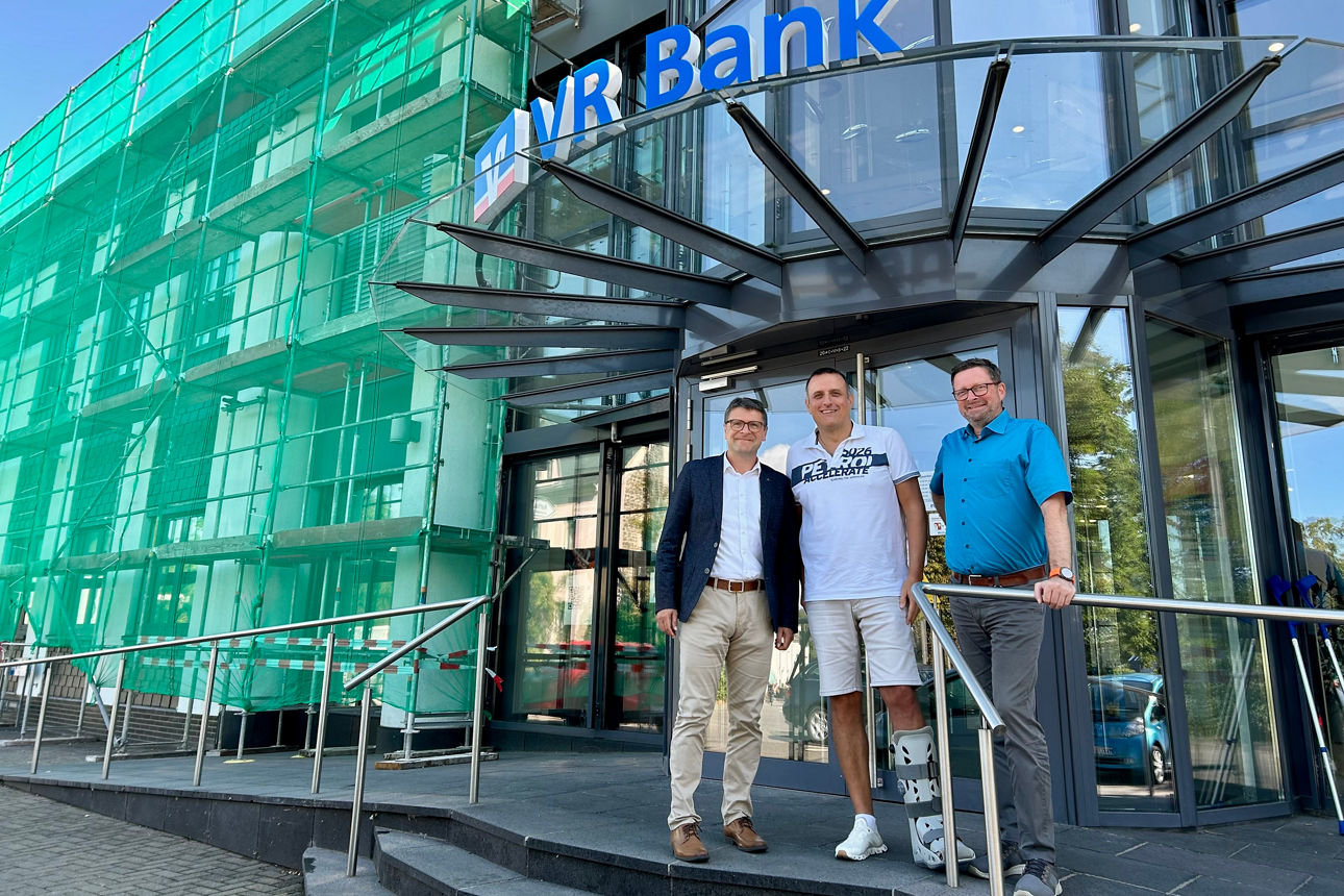 Drei Männer vor VR-Bank-Eingang mit Logoname, Baugerüst links, einer mit Beinschiene, moderner Glasbau, Sommerwetter.