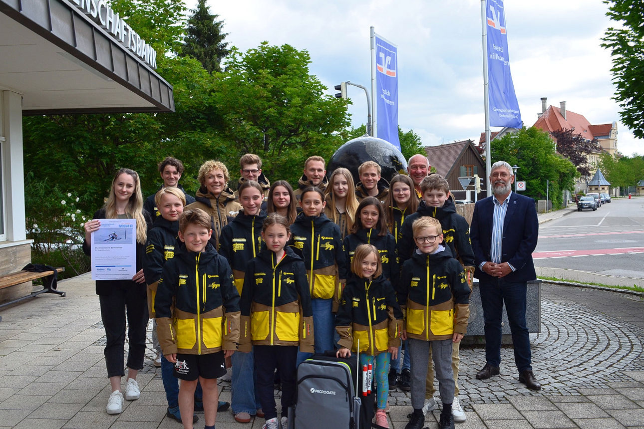 Kinder stehen mit Ihrer einheitlichen Sportkleidung nebeneinander vor unserer Bank mit zwei Mitarbeitern