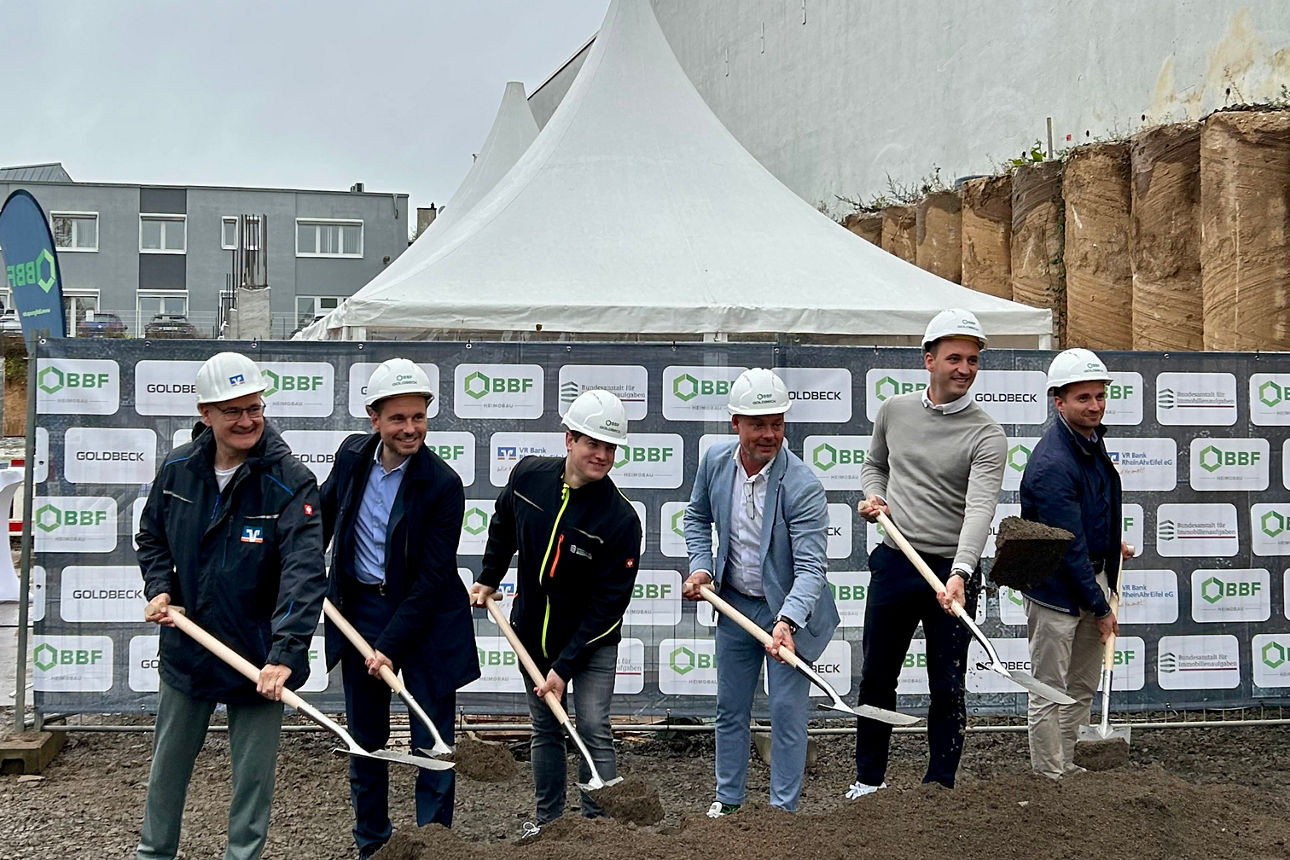 Sechs Personen mit weißen Bauhelmen heben mit Spaten Erde auf einer Baustelle vor Firmenbannern aus.