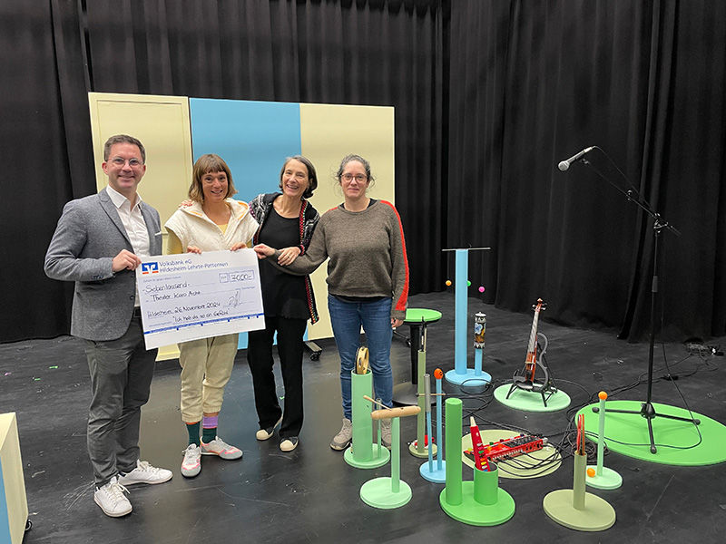 Ein Mann und drei Frauen stehen af einer Bühne neben verschiedenen Musikinstrumenten und halten einen Überreichungsscheck.
