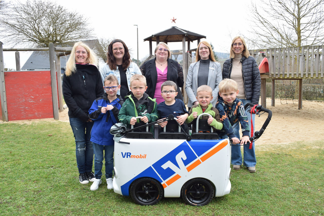 Eine Gruppe von Erwachsenen und Kindern stehen auf einem Spielplatz vor einem Kinderbus zusammen.